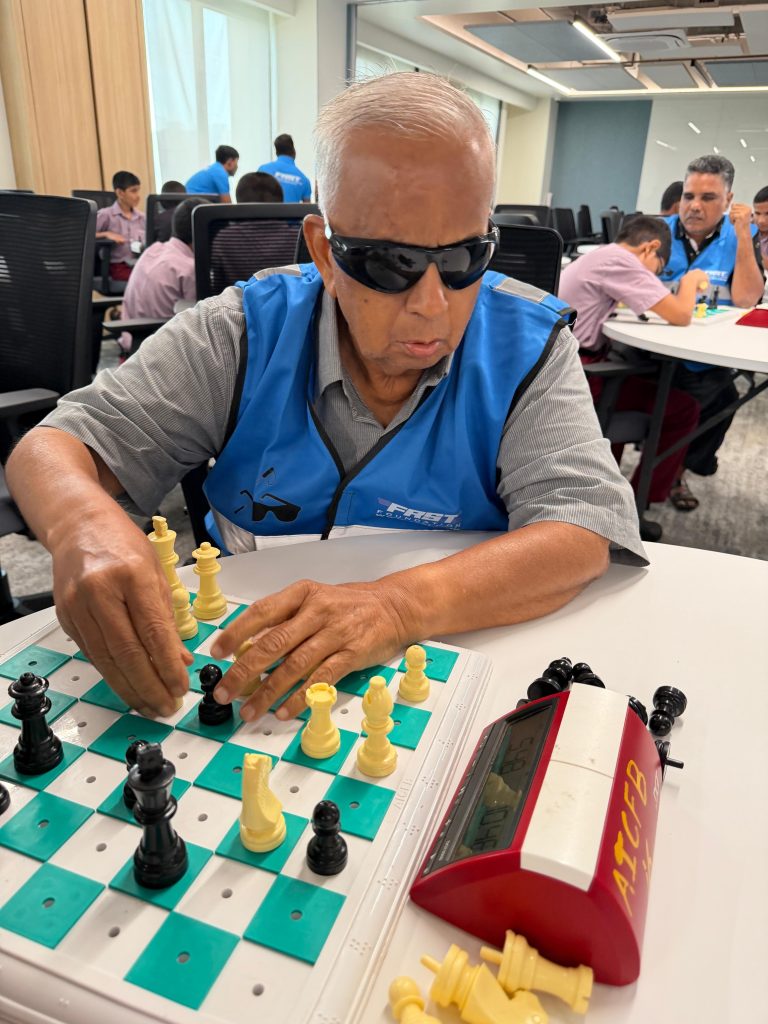 A player reaches across a chessboard during a match, wearing an FRST reflective jacket.