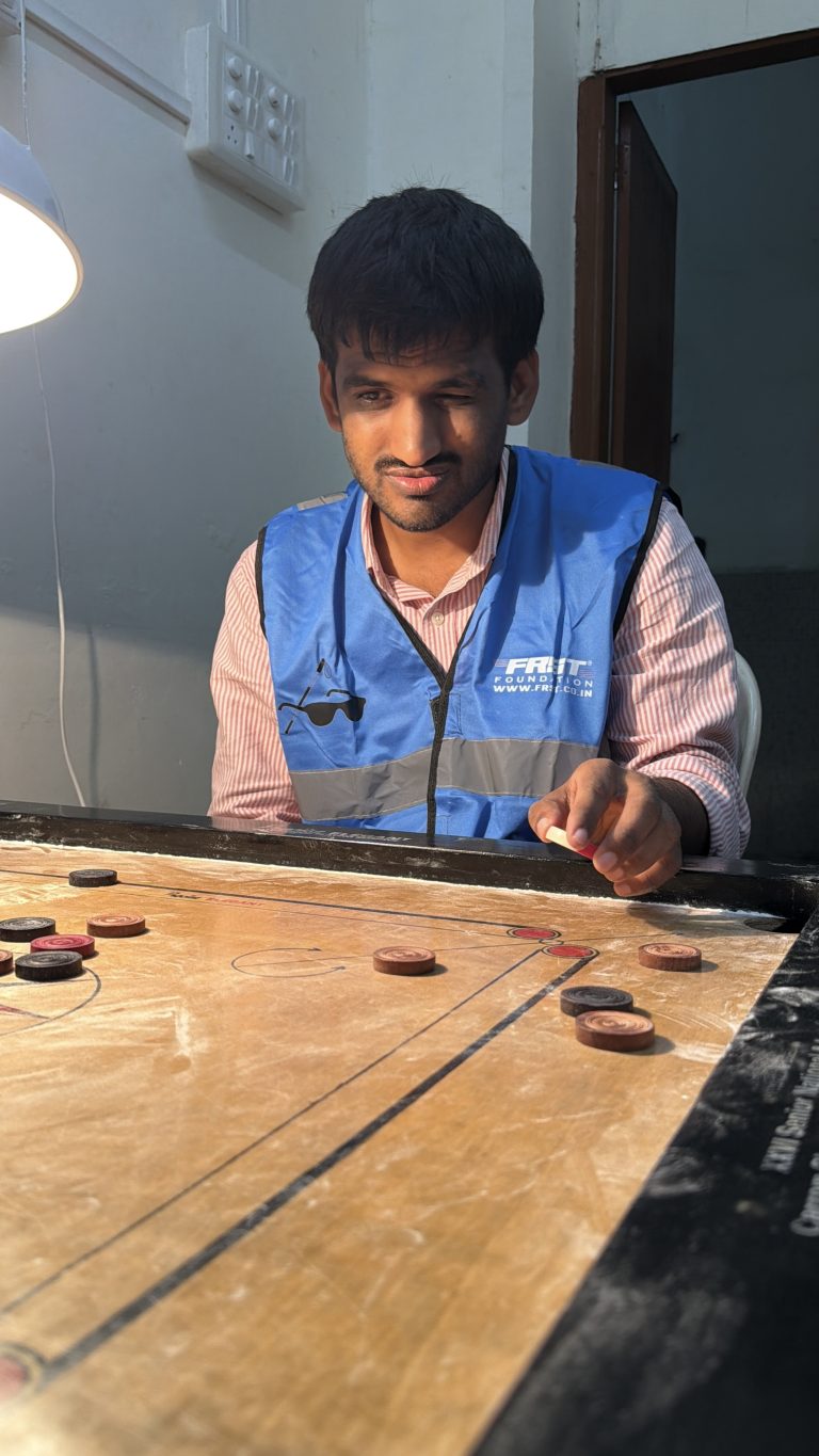 A participant in a blue reflective jacket holds a striker above a carrom board.