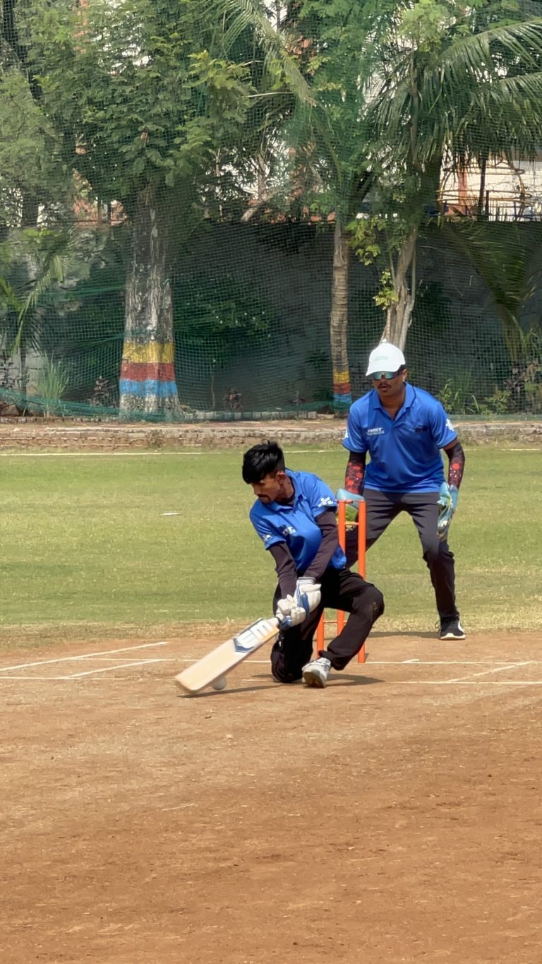 A batter plays a shot while a wicketkeeper waits behind the stumps, both wearing blue FRST shirts.
