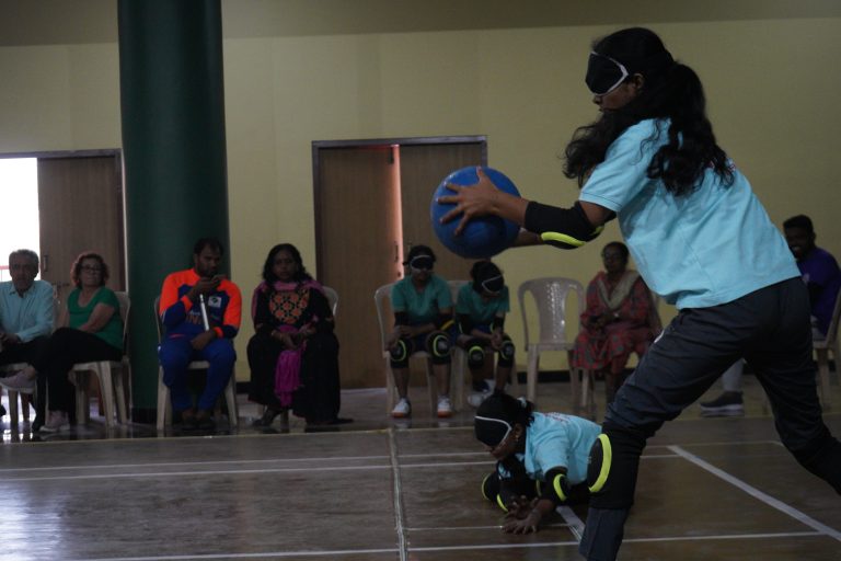 A goalball player wearing eyeshades prepares to throw a blue ball on an indoor court.