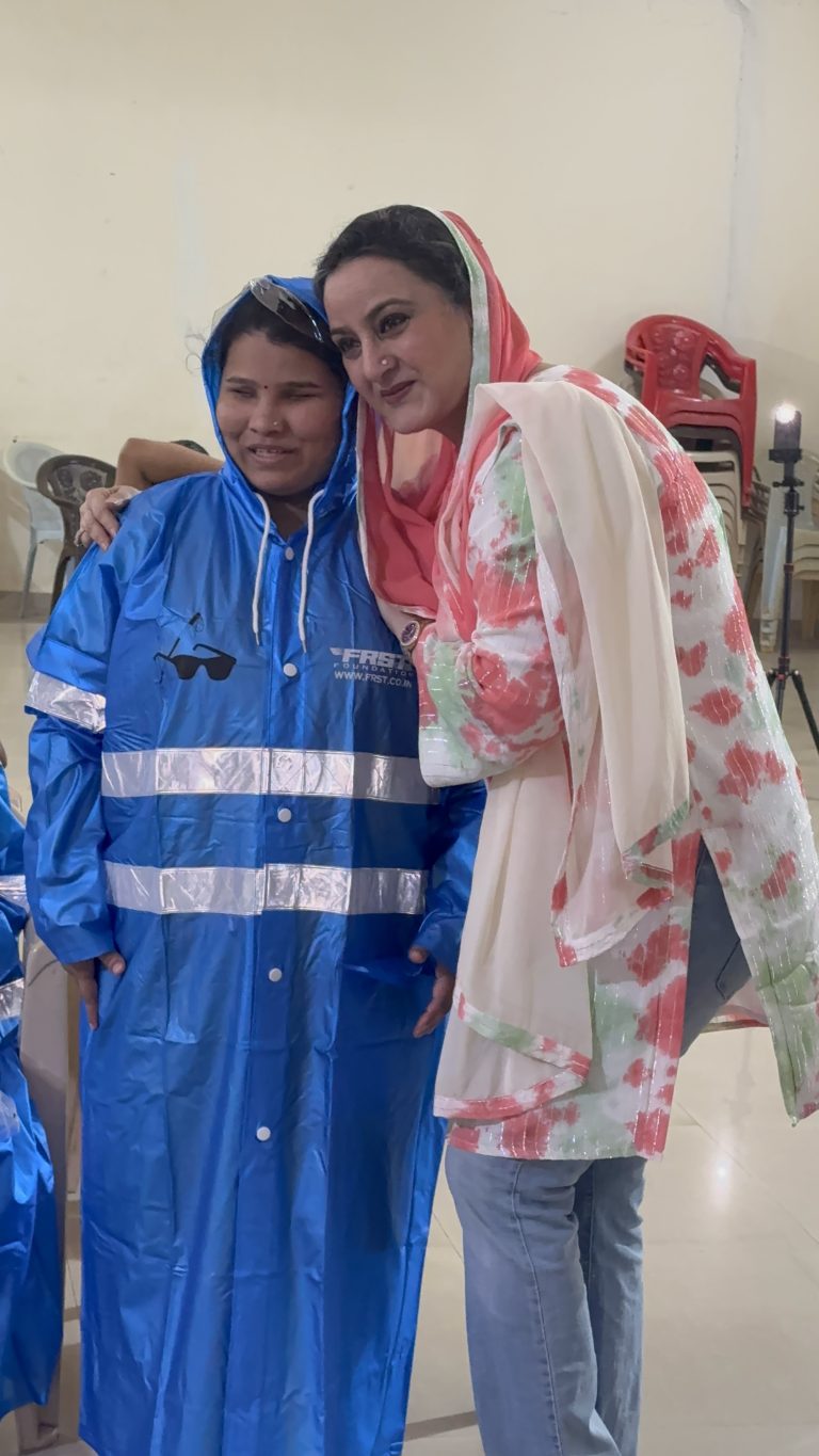 Two women stand together, one wearing a blue reflective FRST raincoat, during the Kumudben visit.