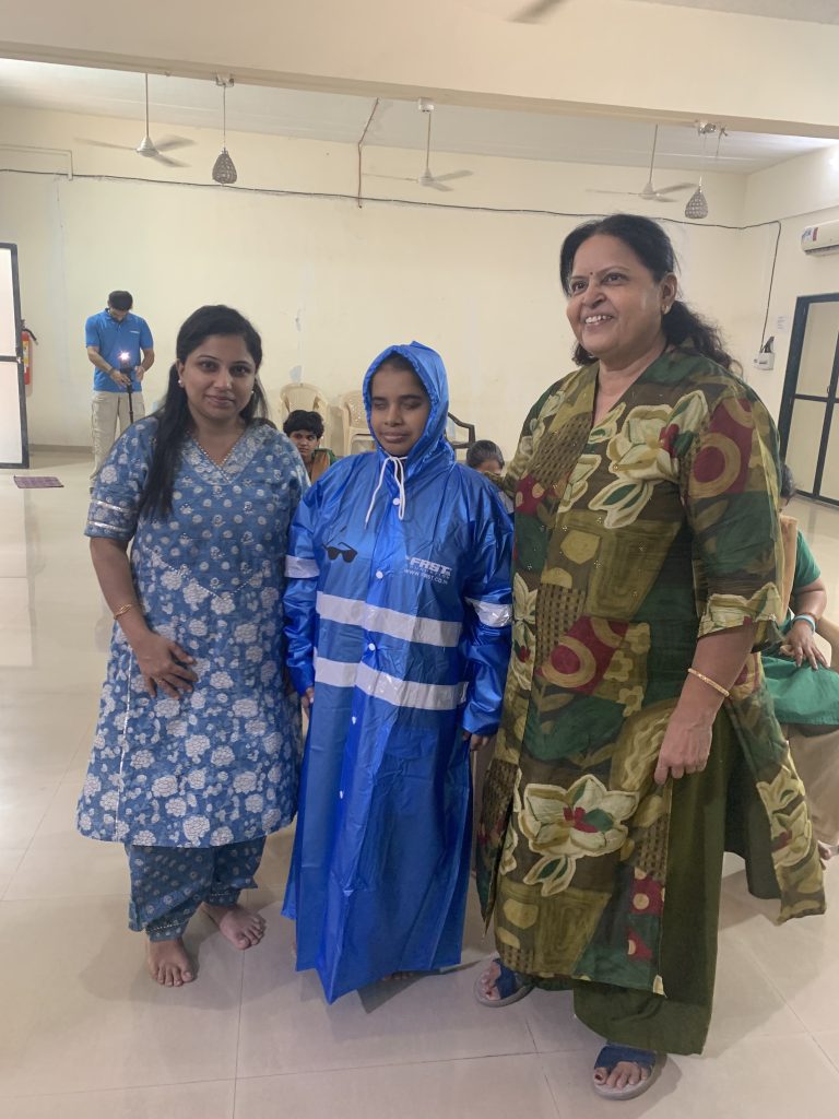 Three women stand together, one wearing a blue reflective FRST raincoat, during the Kumudben visit.